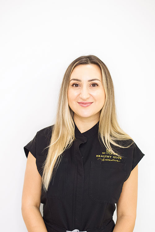 Woman in a black uniform with "Healthy Glow Aesthetics" logo, smiling against a white background.